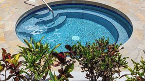 Une piscine circulaire bordée d’une mosaïque, entourée de dalles de pierre et de plantes.