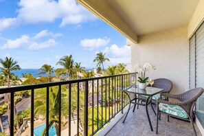 Un balcon avec une table et des chaises, donnant sur une piscine et des palmiers, avec vue sur l’océan.