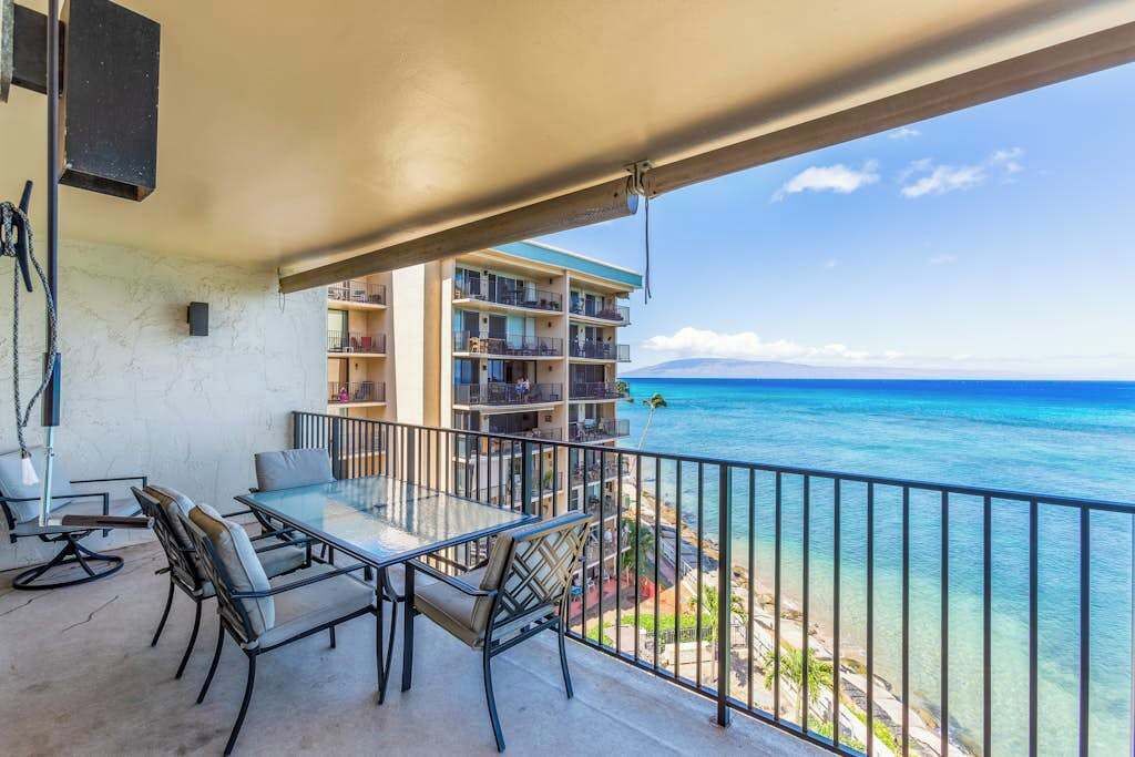 A balcony with a glass table and chairs, overlooking a clear blue ocean and a building with multiple balconies.