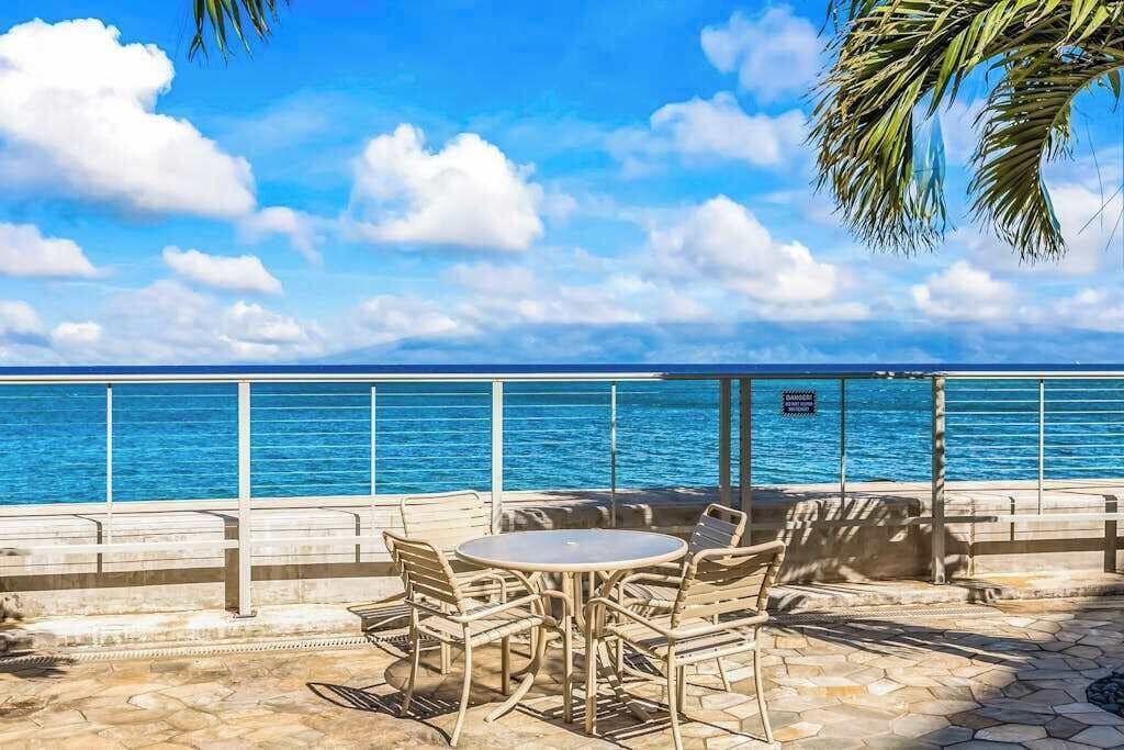 A patio with a round table and chairs, overlooking a body of water with a railing and palm tree.