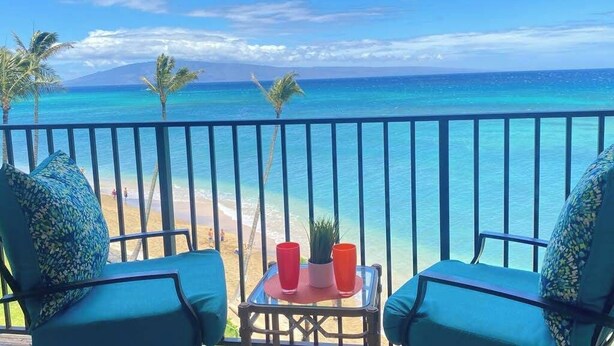 A balcony with two chairs and a small table, overlooking a beach with clear turquoise water and palm trees. - NEW Fall Sale Fifth Floor Kahana Oceanfront with Two Lanais (Lahaina)
