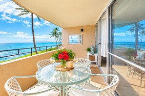 Een tafel met glazen blad en stoelen op een balkon met uitzicht op de oceaan, met palmbomen en de zee.
