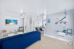 Een moderne woonkamer met een blauwe bank, een eethoek, een keuken en een plafondventilator.