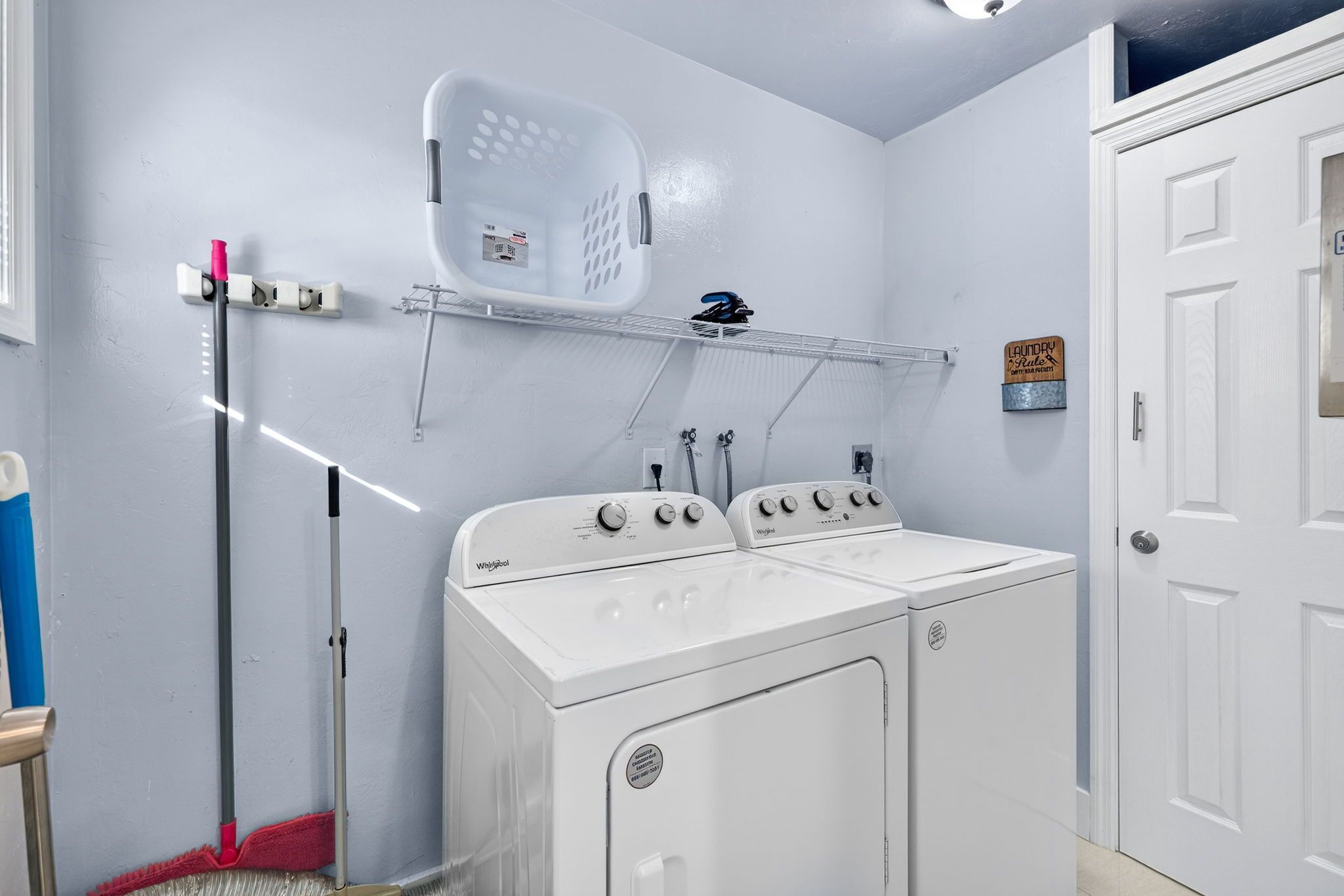 Une buanderie équipée de deux machines à laver et de deux sèche-linge à chargement frontal, tous deux de couleur blanche, ainsi que d’un balai, d’un panier à linge et d’une étagère sur laquelle est accrochée une pancarte.