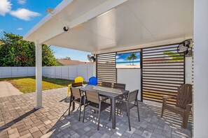 Een overdekt terras met een tafel, stoelen, een ventilator en uitzicht op het strand door een raam.