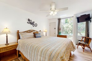 Ein Schlafzimmer mit einem Bett, zwei Nachttischlampen, einem Deckenventilator, einem Fernseher und einem Fenster mit Blick auf Grünflächen.