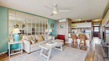 A living room with a sofa, coffee table, and two chairs, a kitchen area with cabinets and a dining table, and a ceiling fan.
