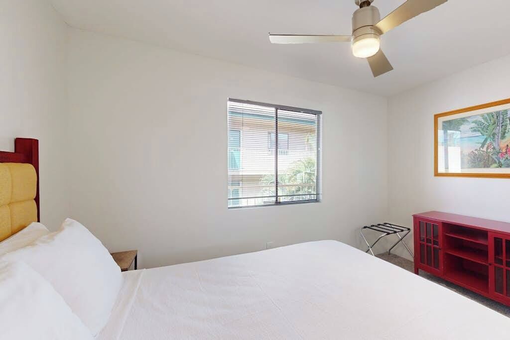 Un dormitorio con cama, ventilador de techo, ventana con persianas, un armario rojo y un cuadro en la pared.