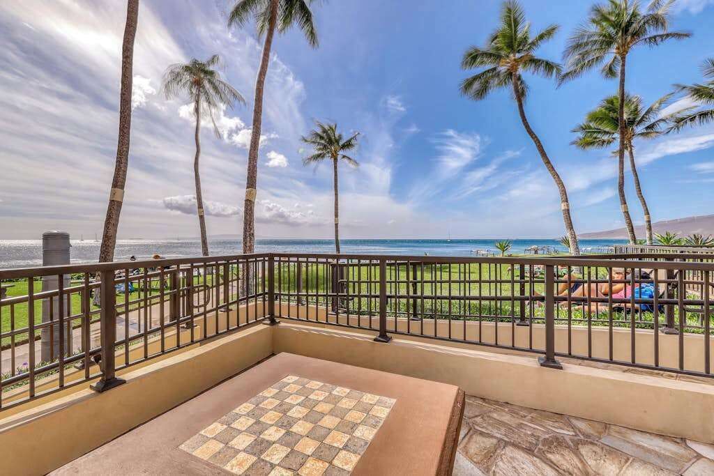 Un balcón con piso a cuadros, barandilla y vista a la playa y palmeras.