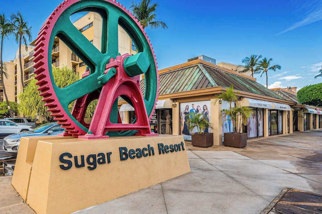 Schild für das Sugar Beach Resort mit einem großen dekorativen Rad, Palmen und einem Gebäude mit Ziegeldach.