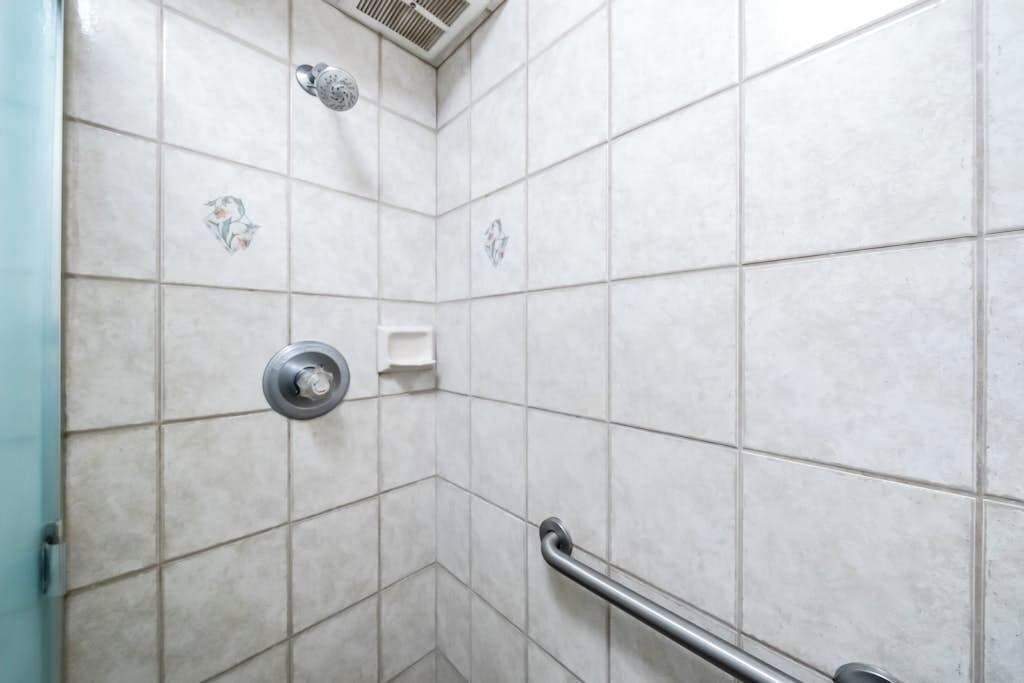 Un baño con azulejos blancos, ducha, barra de apoyo y dispensador de papel higiénico.