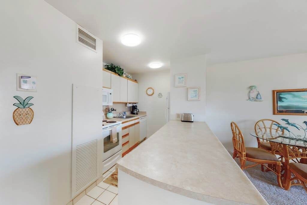 Una cucina moderna con mobili bianchi, un'area pranzo con tavolo in vetro e sedie in vimini e un piccolo frigorifero.