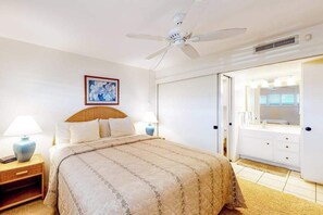 A hotel room with a bed, bedside lamps, a ceiling fan, a bathroom with a mirror, and a door. - New Fall Sale Ocean-View Sugar Beach Penthouse with Pool and Hot Tub (Kihei)