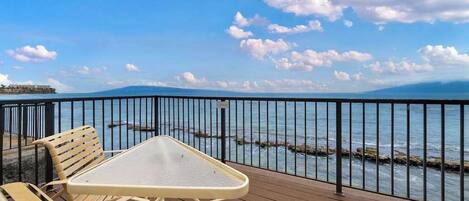 A wooden deck with a table and chairs, overlooking a body of water with a distant shoreline and mountains.