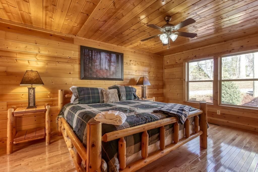 Ein Schlafzimmer einer Holzhütte mit einem Bett, zwei Nachttischlampen, einem Deckenventilator und einem großen Fenster.