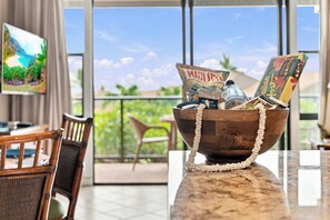 A room with a wooden bowl on a marble table, containing snacks and a bottle, with a view of a balcony and greenery outside. - Beachside Fun at Mahina Surf Resort Condo - Pool, Laundry, Near Local Activities (Lahaina)