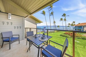 A balcony with a table and chairs, overlooking a lawn and palm trees, with a view of the ocean and a building in the distance. - Lahaina Mahina Surf Villa w/ Pool, Turtle Beach, WD, Near Local Maui Activities (Lahaina)