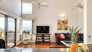 A spacious living room with a large TV, ceiling fans, a couch, and a dining area with a fruit bowl. - NEW Fall Sale Ocean-View Wailea Townhouse with Two Shaded Lanais (Wailea)