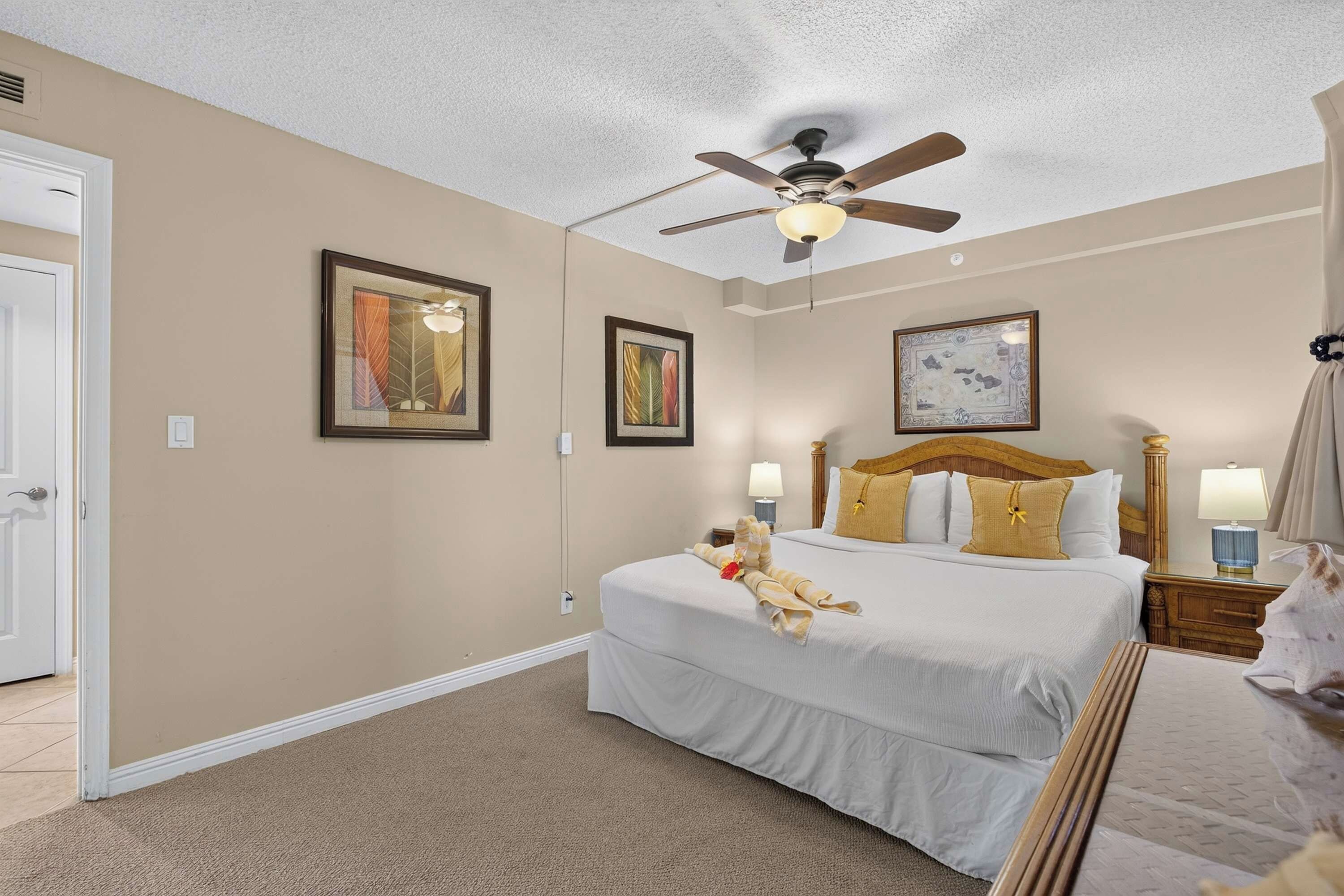 A hotel room with a bed, a ceiling fan, two bedside lamps, and framed artwork on the walls.