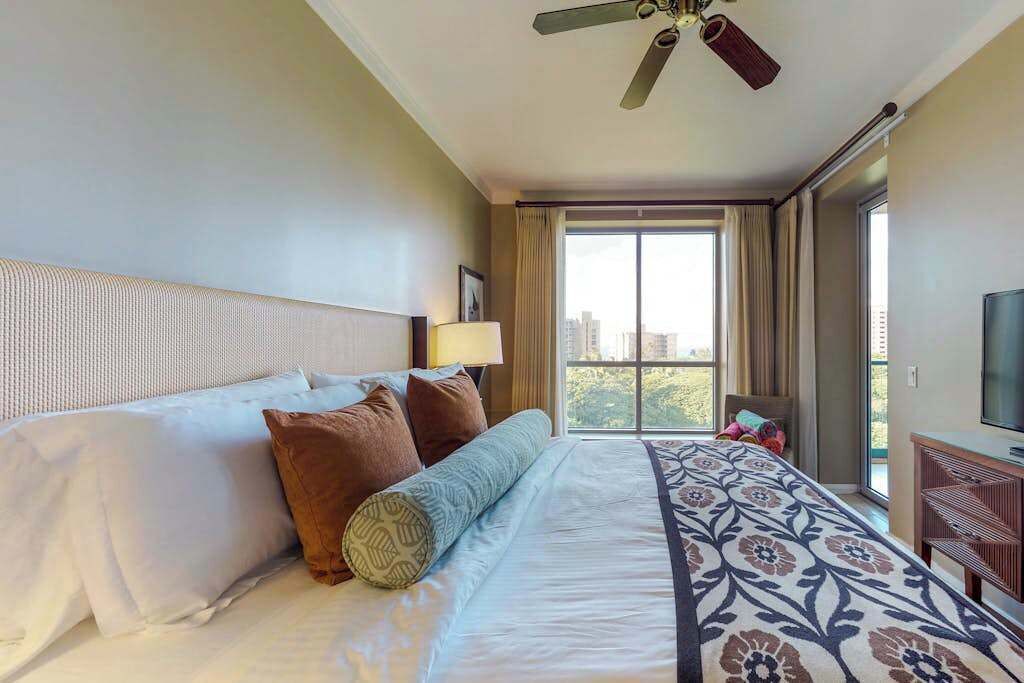A hotel room with a large bed, a ceiling fan, a TV, and a window with curtains.