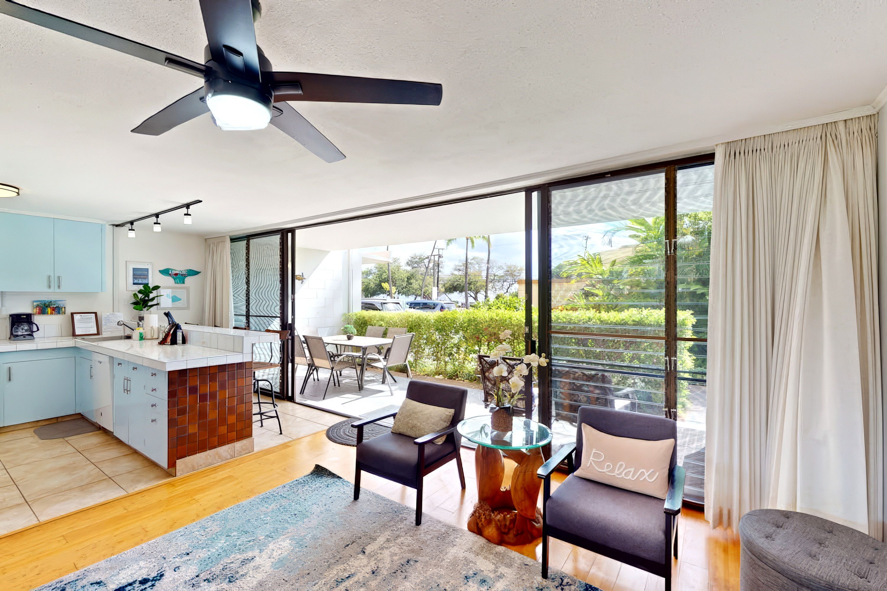 A modern kitchen with a central island, a dining area with a glass table and chairs, and a balcony with a view of greenery.