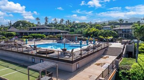 A swimming pool with lounge chairs and umbrellas, surrounded by a fence, with buildings and palm trees in the background. - NEW Kaanapali Studio with Golf and Ocean Views Near the Beach (Lahaina)