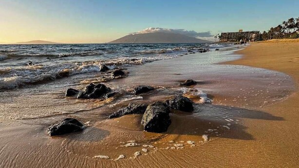 Une plage avec des vagues, des rochers et une montagne qui se profile à l’horizon.