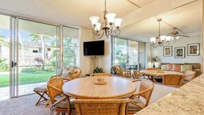 A spacious living area with a large sliding glass door leading to a patio, a round wooden table with wicker chairs, a sofa with cushions, and a ceiling fan. - *New* Fall Special Garden Lanai Retreat Near Kamaole II (Kihei)