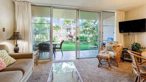 A living room with a sofa, coffee table, and chairs, a television mounted on the wall, and a sliding glass door leading to an outdoor patio with a table and chairs. - *New* Fall Special Garden Lanai Retreat Near Kamaole II (Kihei)