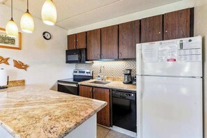 A kitchen with granite countertops, dark wooden cabinets, a white refrigerator, a microwave, a coffee maker, and a clock on the wall. - *New* Fall Special Garden Lanai Retreat Near Kamaole II (Kihei)