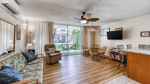 Una sala de estar con sofá, sillones, una mesa de comedor y sillas. Hay un televisor montado en la pared, un ventilador de techo y un gran ventanal con cortinas.