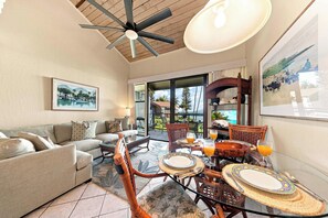 A spacious living area with a sofa, dining table, and chairs, a ceiling fan, and a large window offering a view of the outdoors. - Mahina Surf Oceanfront Resort Condo-Pool, BBQ, Kitchen, Lanai, Sea Turtle Beach (Lahaina)