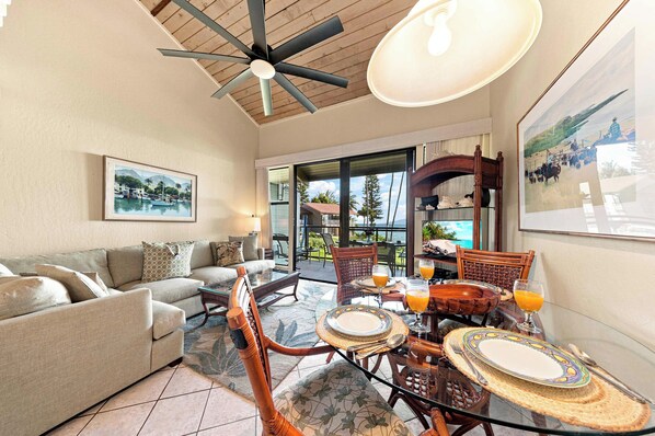 A spacious living area with a sofa, dining table, and chairs, a ceiling fan, and a large window offering a view of the outdoors. - Mahina Surf Oceanfront Resort Condo-Pool, BBQ, Kitchen, Lanai, Sea Turtle Beach (Lahaina)