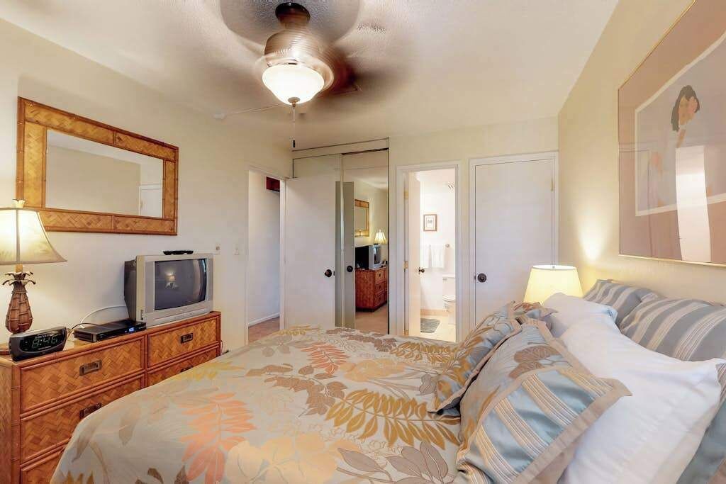 A bedroom with a bed, a television on a wooden dresser, a ceiling fan, and a door leading to another room.