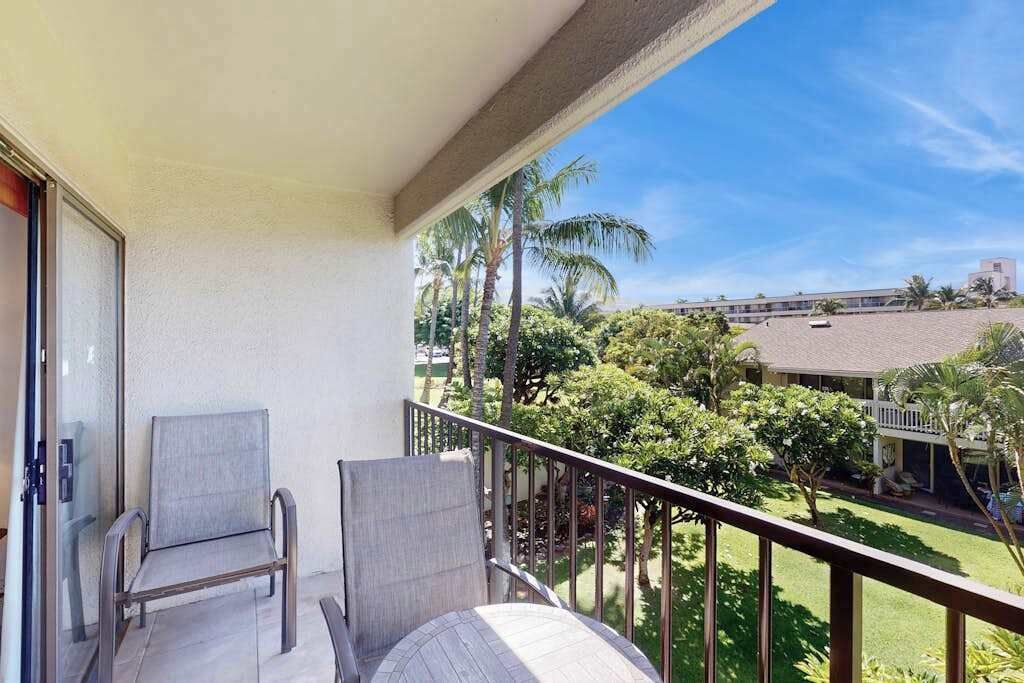 Ein Balkon mit zwei Stühlen und einem Tisch, mit Blick auf eine grüne Landschaft mit Palmen und einem Gebäude im Hintergrund.