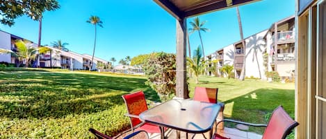 A covered outdoor seating area with a table and chairs, surrounded by a well-maintained lawn and palm trees, with buildings in the background.