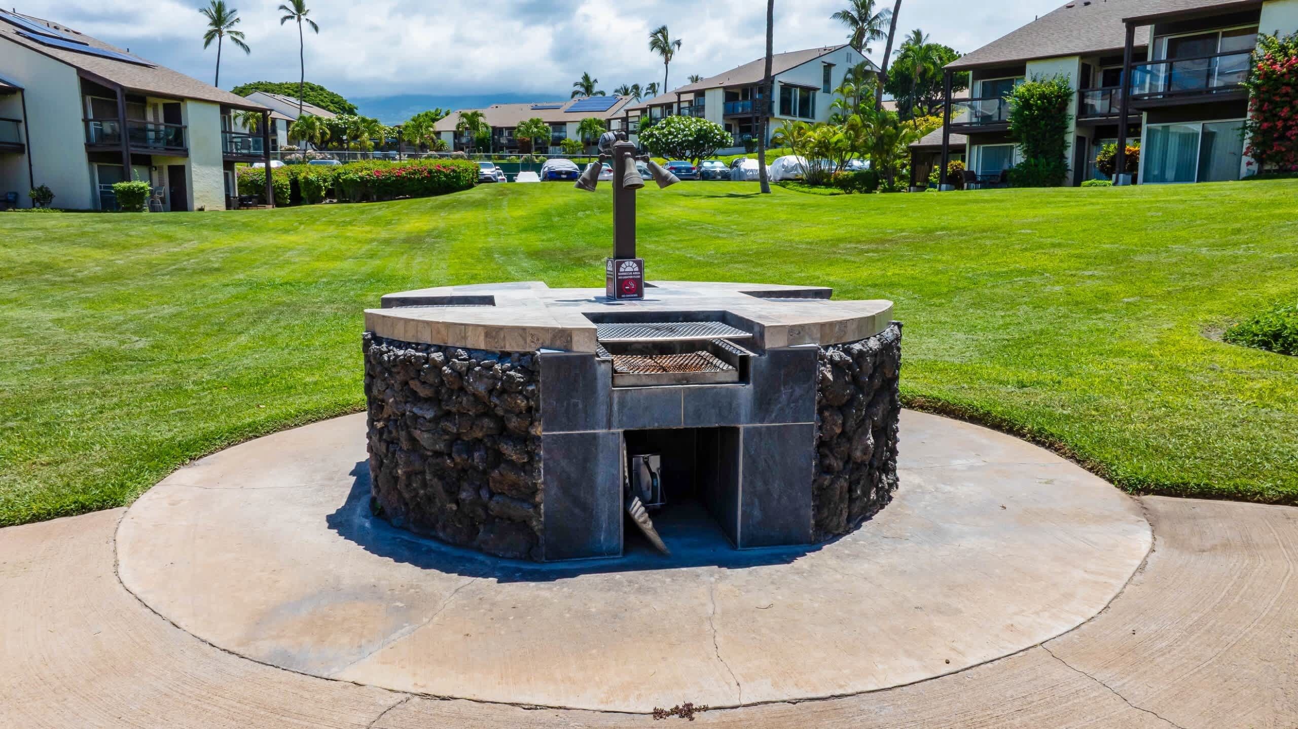 Un espacio de estar al aire libre con forma circular, que incluye una parrilla de piedra y metal, rodeado por un sendero pavimentado.