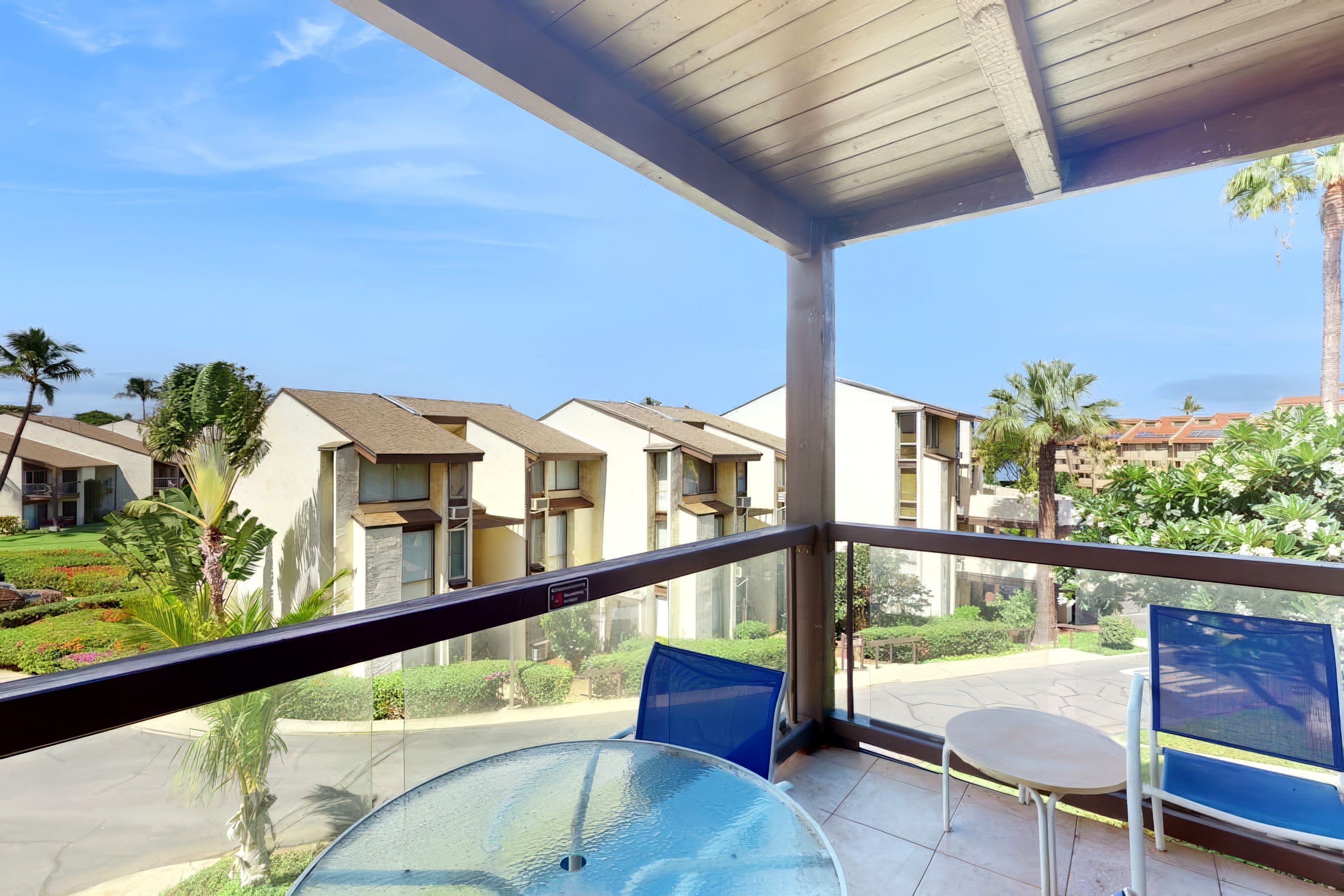 A balcony with a glass table and chairs, overlooking a residential area with multiple buildings and palm trees.