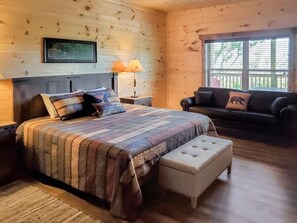 A wooden cabin bedroom with a bed, nightstand, lamp, sofa, and window. - Smoky Mountain Dreams (Gatlinburg)