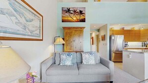 Ein Wohnzimmer mit grauem Sofa, einem Holzschrank und einer Küche mit Holzausstattung und einem Kühlschrank.