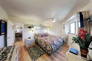 Ein Schlafzimmer mit Bett, Nachttischlampen, Deckenventilator, Fenster mit Vorhängen, einer Pflanze und einem Fernseher an der Wand.