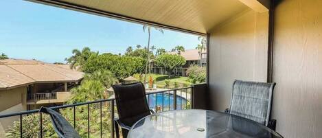 Un balcón con mesa y sillas de vidrio, una piscina y vistas a otros edificios y zonas verdes.