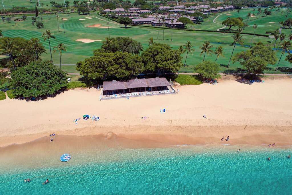 Une vue aérienne d’une plage avec un bâtiment, des palmiers et un terrain de golf en arrière-plan.
