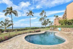 A swimming pool with a curved edge, surrounded by a stone deck, palm trees, and a view of the ocean. - New 70 Feet from Ma'alaea Bay, Pool and Hot Tub (Wailuku)