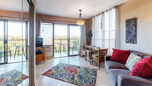 Een ruime woonkamer met een bank, televisie, eettafel en stoelen, grote ramen en een balkon.