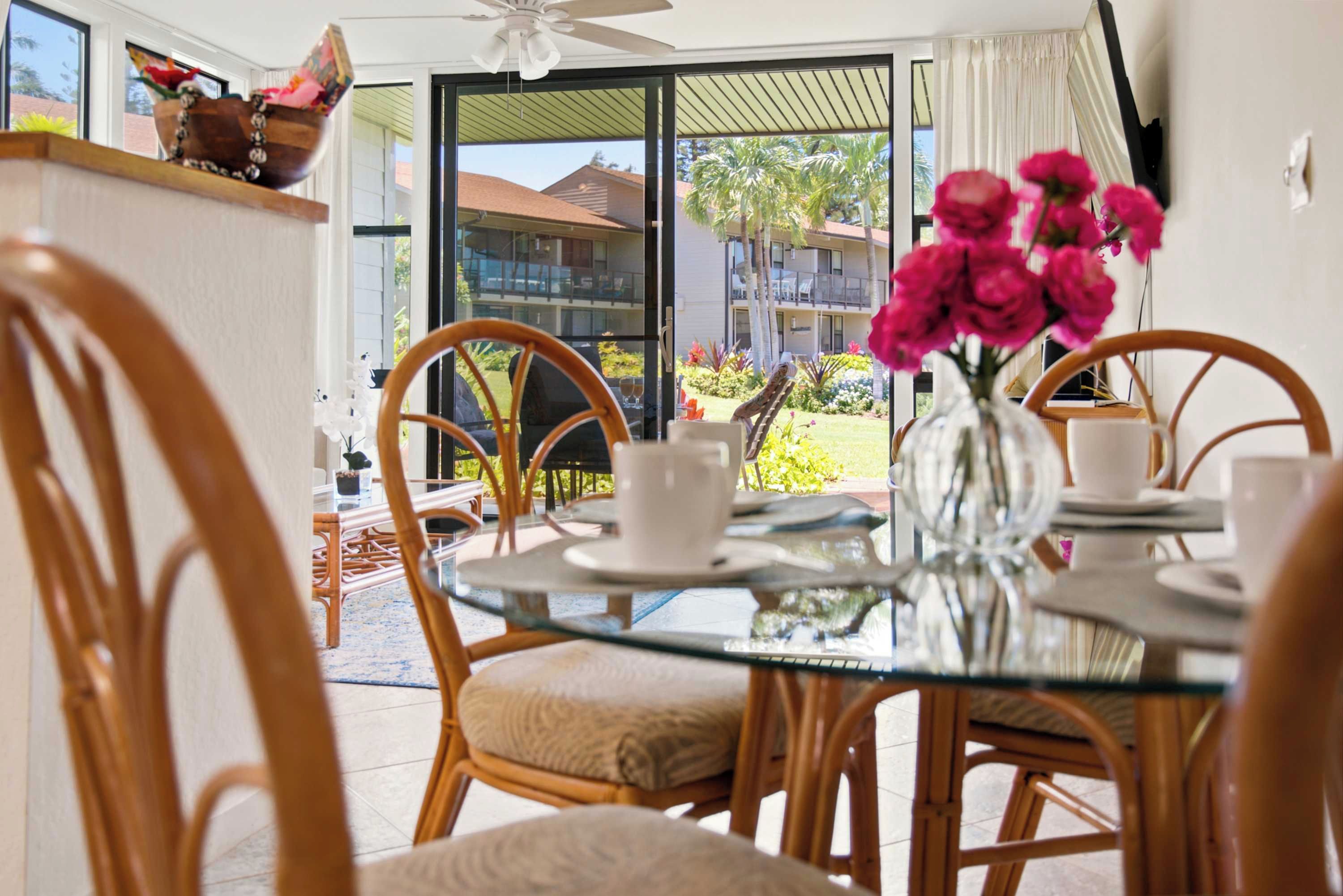 Espaço de refeições com mesa de vidro, cadeiras de vime e vista para um edifício e jardim.