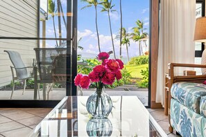 A room with a glass door leading to a view of palm trees and a lawn. There is a chair, a sofa, and a table with a vase of flowers. - Ground Floor Mahina Surf Oceanfront Condo at Turtle Beach- Pool, Kitchen, WD, TV (Lahaina)