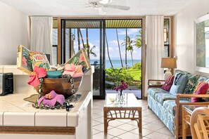 A room with a view of the ocean, a wooden coffee table, a sofa, and a dining table with a vase of flowers. - Ground Floor Mahina Surf Oceanfront Condo at Turtle Beach- Pool, Kitchen, WD, TV (Lahaina)