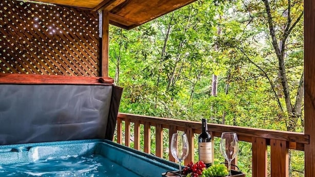A hot tub with a wooden deck and a wooden railing, surrounded by lush greenery. - The Blessing Cabin (Sevierville)