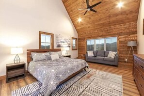 A bedroom with a bed, a gray sofa, a wooden ceiling, and a large window. - Top of the Summit Lodge (Sevierville)